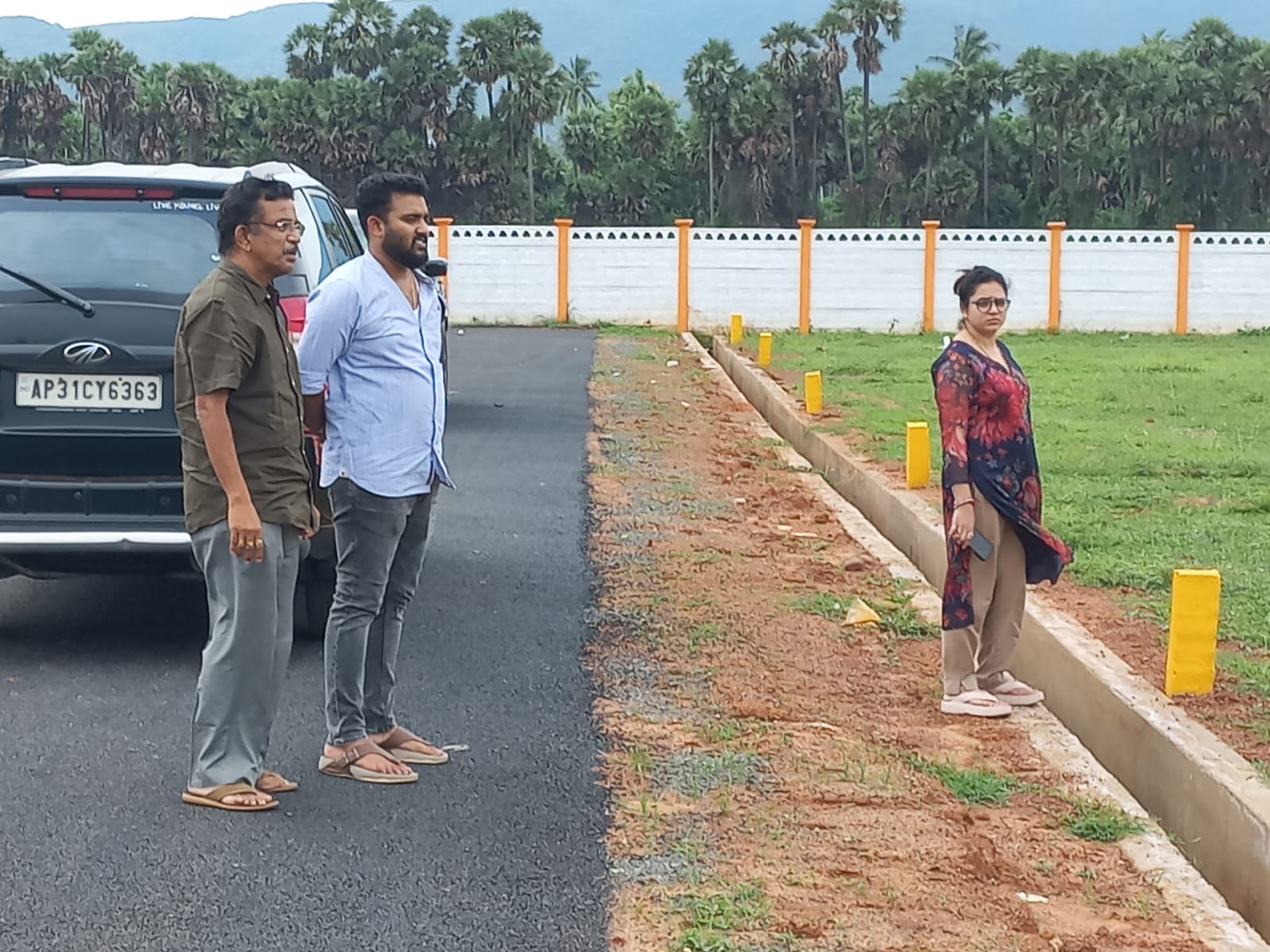 Blacktop internal road and white compound wall at the Adhvik Aero City venture, with plot markers, palm trees and the hills behind